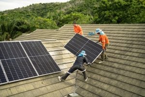 Three workers in hats and orange shirts install solar panels in East Baton Rouge Parish on a sloped, shingle roof, with lush trees and greenery in the background.