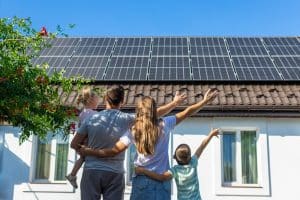 A family of four stands outside their house, proudly pointing at solar panels installed by St. John the Baptist Parish Solar Installers under a clear blue sky.
