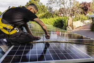A person from St. John the Baptist Parish Solar Installers, wearing safety gear, installs or maintains solar panels on a rooftop with a screwdriver on a sunny day.