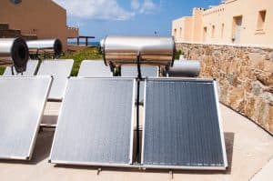 Rows of Solar Water Heater systems with metal tanks and solar panels are installed outdoors near a stone wall and beige buildings under a partly cloudy sky.