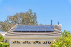 Solar panels are installed on the roof of a residential garage, surrounded by trees and plants, under a clear blue sky—a perfect example of solar in California.